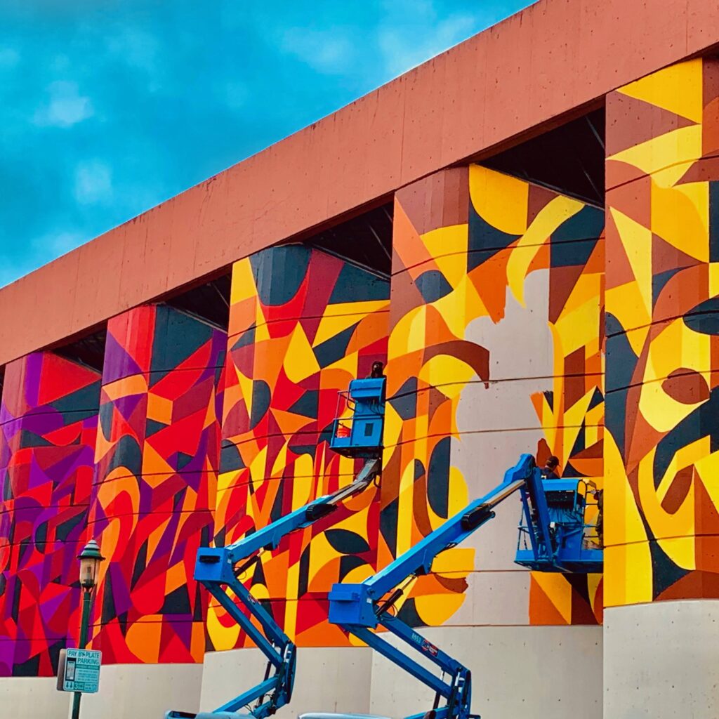 The face of the Baltimore County Library where the mural is being painted. Of the six panels of wall being painted, the first through fourth are complete. The artists are standing on two separate boom lifts painting the fifth wall panel. The sixth panel is complete. The mural has a gradient color scheme, more purple on the far left panel, progressing to red, to orange, to yellow on the far right panel.